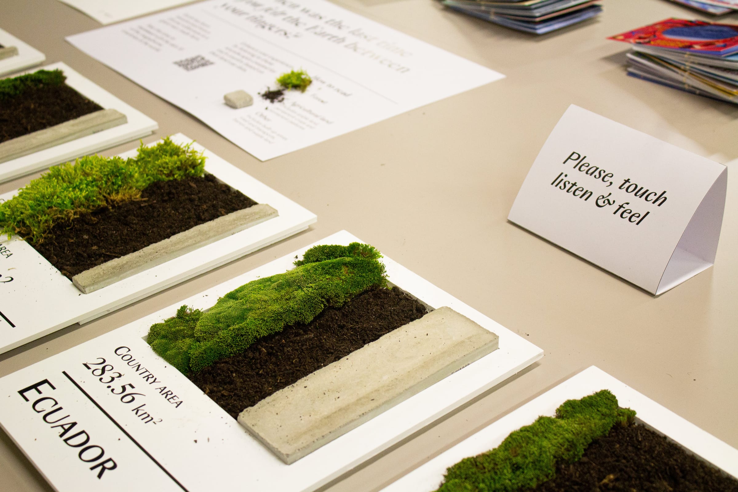 Moss, soil and concrete boards on a table next to a card that reads “Please, touch, listen & feel”.